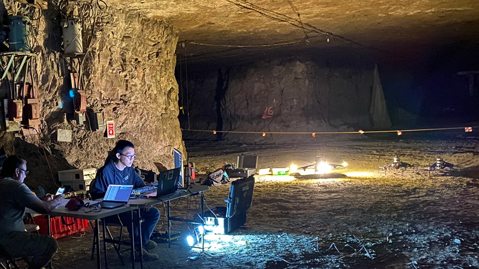 Researchers work at laptops inside a dim underground cave, surrounded by rugged rock walls, cables, and bright equipment lights.