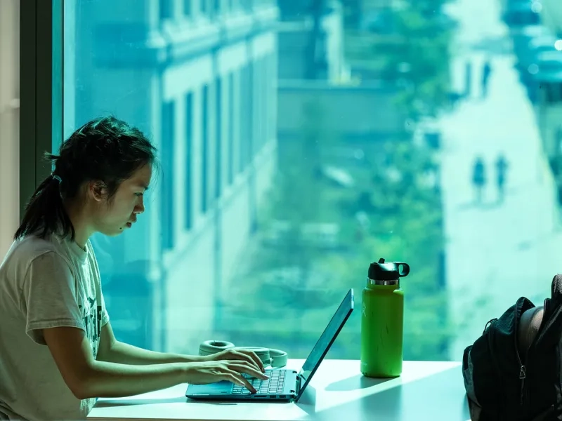 Student at a table working on a computer in front of window with view of campus outside