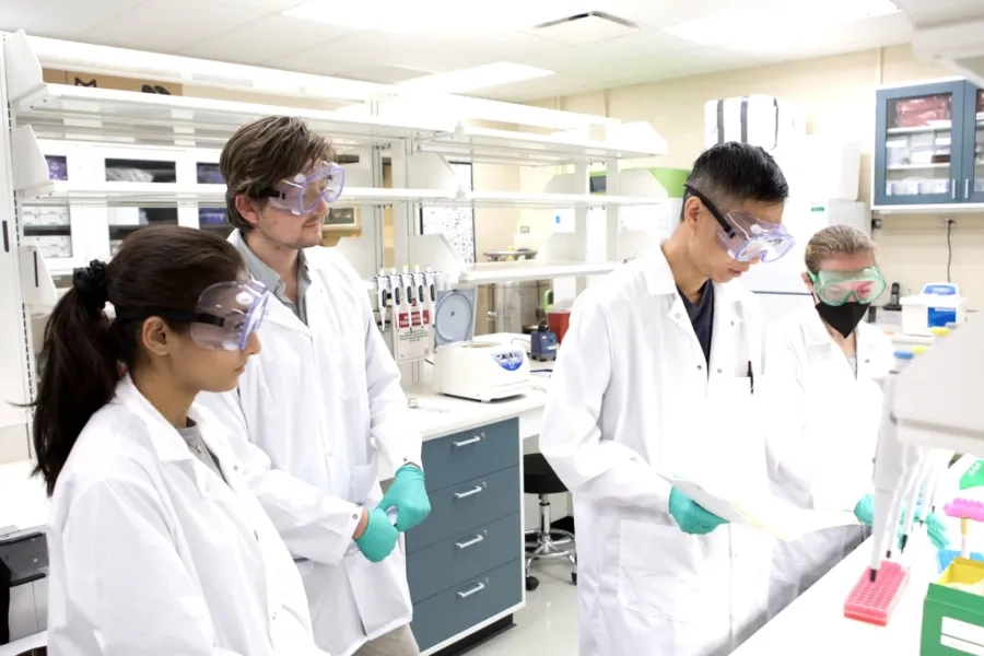 Four CMU students in lab coats and safety goggles work on a research project, standing in front of equipment in a laboratory